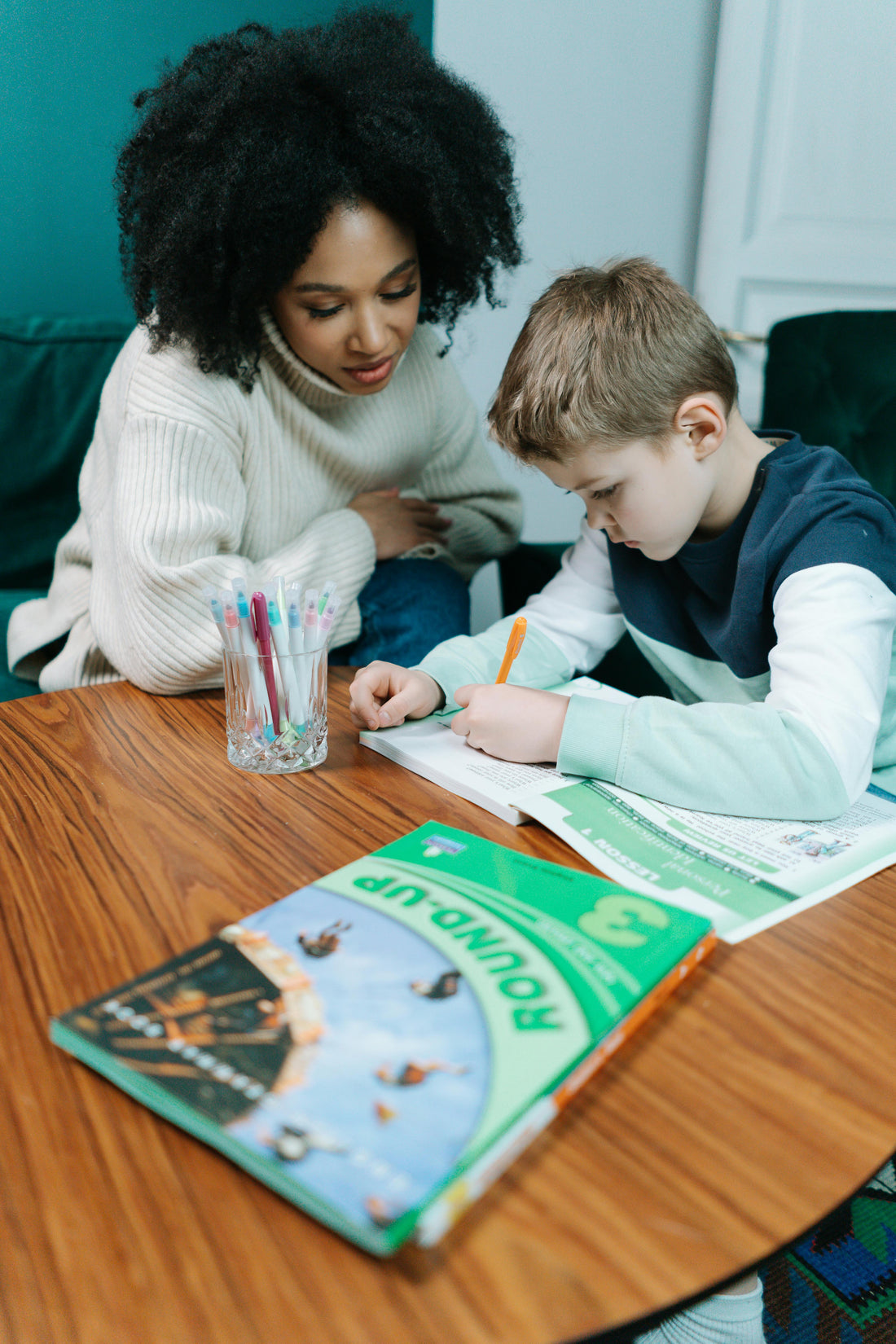 A woman with tight black curls and a sweater sits at a wooden table with a young kid with short hair working on their homework.